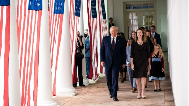 Trump e Amy caminham em corredor da Casa Branca, com outras pessoas atrás e bandeiras dos EUA ao lado