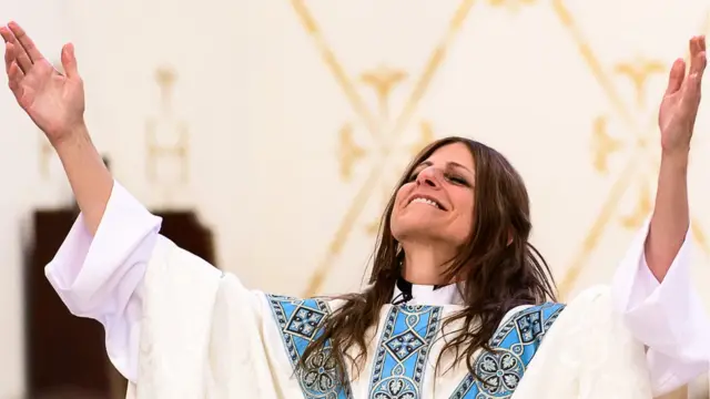 Fotografia colorida mostra mulher branca de cabelo comprido sorrindo com as mãos para o alto e usando roupas de padre