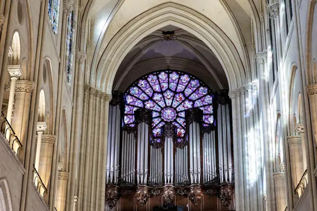 Uma vista interna da catedral mostra janelas e vigas ornamentadas, com um vitral roxo na frente, acima dos muitos tubos prateados do órgão restaurado