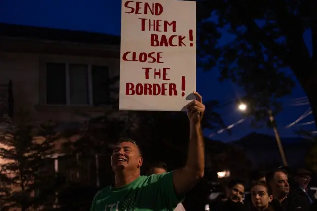 Moradores locais e ativistas contra migrantes se reúnem para o oitavo protesto contra o uso da escola fechada St. John Villa Academy, em Staten Island, Nova York, como abrigo para migrantes. Um homem em uma camiseta verde segura uma placa com a frase: "Mandem-nos de volta! Fechem a fronteira!"