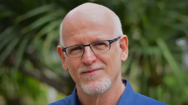 Daniel Becker é um homem de pele clara, careca, com cavanhaque e bigode brancos. Está usando um óculos retangular de aro escuro e uma camisa azul. 