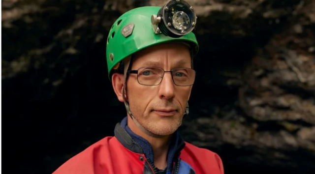 Phil Short com capacete verde e lanterna frontal, vestindo roupa de exploração, olhando diretamente para a câmera. Fundo escuro, possivelmente na entrada de uma caverna.