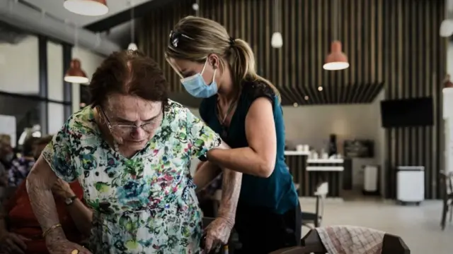 Um voluntário ajuda a apoiar uma mulher em pé acima de uma cadeira em uma mesa durante o almoço no site Landais Alzheimer para pacientes com Alzheimer em Dax, França