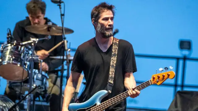 Keanu Reevers de camiseta preta aparece tocando guitarra em uma apresentação. Ao fundo, um baterista tocando bateria