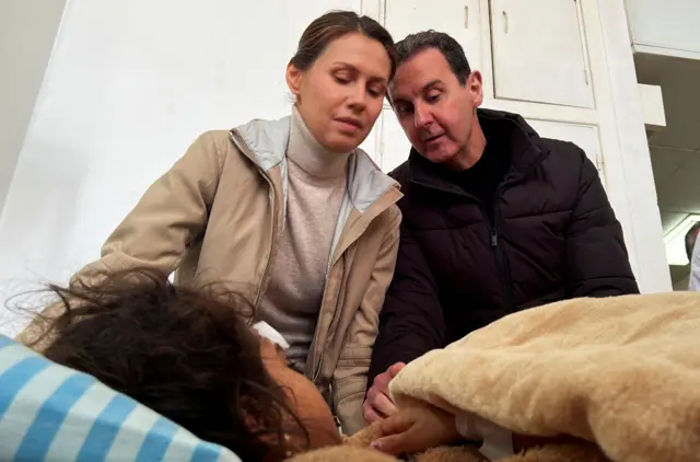 O ex-presidente sírio Bashar al-Assad e sua esposa Asma visitam uma sobrevivente do terremoto no Hospital Tishreen em Latakia, Síria, após um terremoto. Eles são vistos olhando para uma menina quase totalmente coberta por um cobertor em uma cama de hospital. Asma está vestindo uma jaqueta bege com uma gola rolê bege, enquanto Bashar al-Assad está com uma jaqueta preta sobre uma camiseta preta.