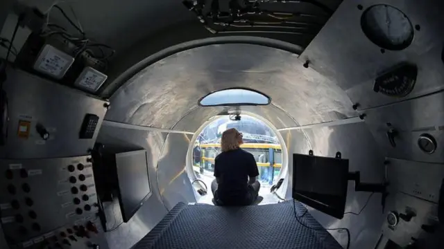 Vista interna do submarino, com mulher sentada de costas na ponta