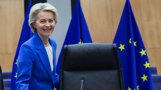 A presidente da Comissão Europeia, Ursula Von der Leyen, uma mulher branca de cabelo curto loiro claro ficando grisalho, com um blazer  azul, em frente a três bandeiras da união europeia