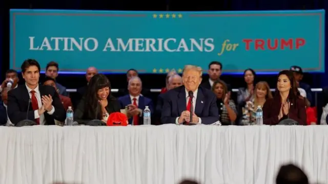 Trump no meio de mesa, sentado ao lado de várias pessoas sorrindo; ao fundo, telão diz 'Latino-americanos por Trump'