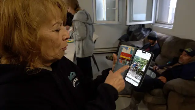 Uma senhora de pele e cabelos claros segura o celular e mostra uma foto para a câmera. No fundo, dois homens sentados em um sofá. 