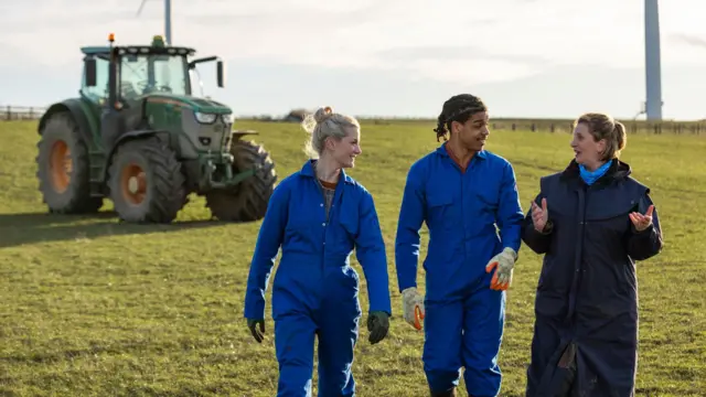 Dois jovens de macacão ao lado de profissional mais experiente da área de agricultura, com trator ao fundo.