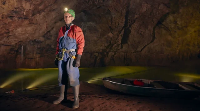 Phil Sort usando capacete com lanterna e roupa de exploração em uma caverna. Ele está ao lado de um pequeno barco.