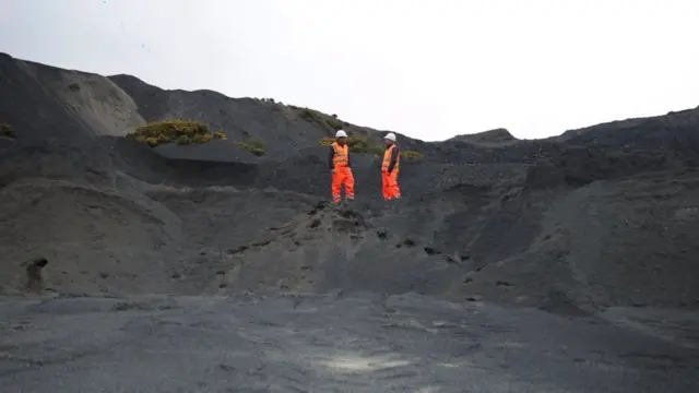 Dois homens de uniforme laranja conversam entre montes de rocha negra