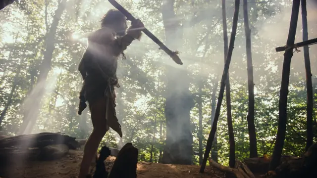 Uma recriação de um homem pré-histórico com sua lança em posição de combate na floresta.