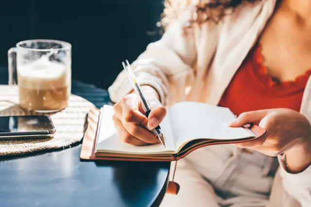 Uma pessoa anota coisas em uma caderno, apoiado sobre uma mesa onde há uma xícara de café e um telefone celular.