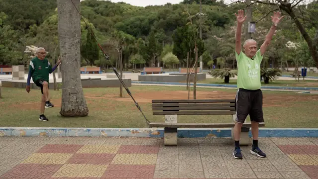 Dois homens idosos com cabelos brancos se alongam em parque, com muitas árvores ao fundo.