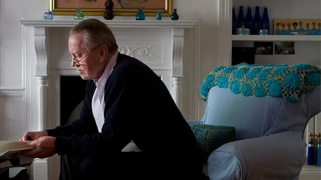 Chuck Feeney sentado de perfil em sala de estar, olhando para livro