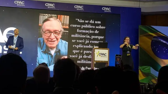 Eduardo Bolsonaro fala no palco, com imagem e frase de Olavo de Carvalho na tela atrás