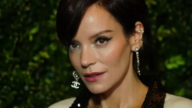 Lily Allen, fotografada com cabelos pretos em um penteado para cima com franja lateral, delineador de olhos e brincos de diamante