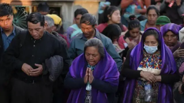 Na foto, um homem do lado esquerdo vestido de preto com as mãos unidas olha para baixo. Ao ldo dele, uma mulher com as mãos em posição de oração, vestida com um pano roxo sobre a roupa, ao lado, uma mulher de máscara, com um pano roxo igual sobre a cabeça. No fundo, várias pesssoas