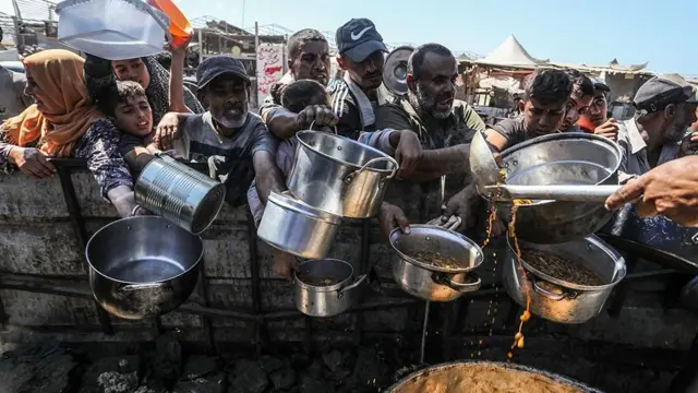 Homens, mulheres e crianças estendem panelas atrás de muro pedindo comida que está sendo distribuída