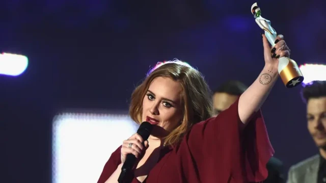 Adele fala no microfone enquanto segura um troféu