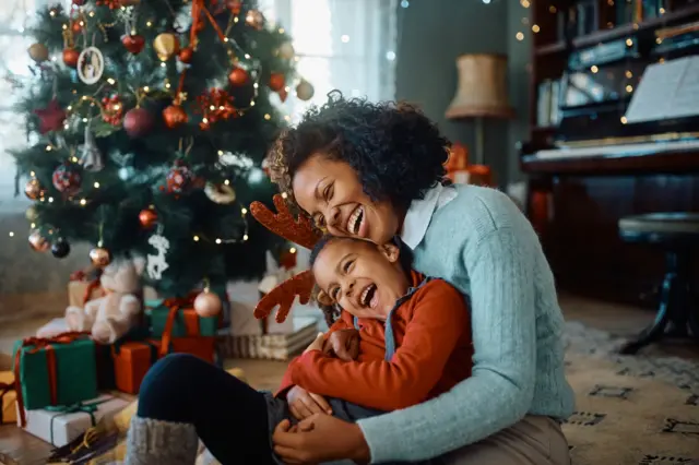 Mãe e filha rindo abraçadas sentadas no chão em frente a uma árvore de Natal rodeada de presentes