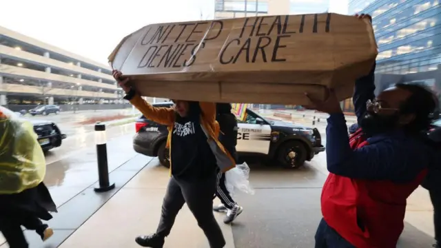 Duas pessoas no meio de um protesto segurando um caixão com a inscrição "United Health nega atendimento"