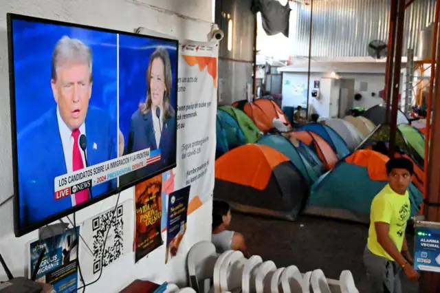 Debate passando na TV, com pessoas e barracas de acampamento dentro de abrigo