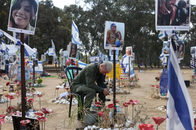 Um homem sentado em uma cadeira no memorial onde ocorreu o festival de música — ele está cercado por bandeiras israelenses e fotos de pessoas que foram mortas no ataque.