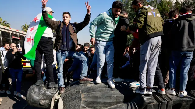 Un groupe de jeunes manifestants, dont l'un est drapé dans le drapeau de groupes rebelles syriens, posent pour les caméras alors qu'ils piétinent le visage d'une statue représentant un parent du président syrien déchu.