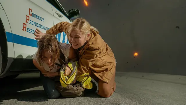 As atrizes Kirsten Dunst e Cailee Spaeny ajoelhadas ao lado de uma viatura de polícia, se protegendo da queda de detritos, em cena do filme 'Guerra Civil' 