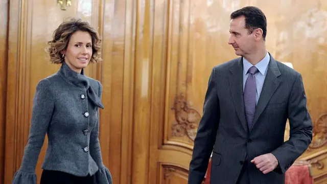 Asma al-Assad, who has short curly hair, wears a grey jacket and looks off to the side while her husband Bashar, wearing a dark grey suit and tie, looks at her. They are stood in a wood-panelled room with red-cushioned seating in the background. This is a stock photo from 2010.