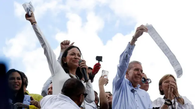 María Corina Machado e Edmundo Gonzalez erguem os braços, rodeados por outras pessoas, em cima de carro de som