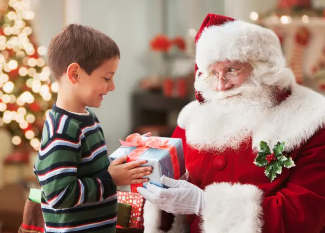 Menino recebendo presente de homem vestido de Papai Noel com decoração de Natal ao fundo