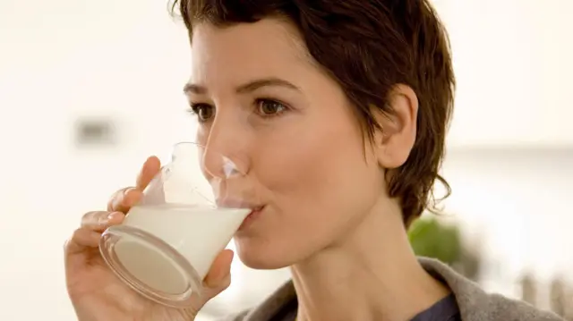 Une femme aux cheveux bruns courts boit du lait dans un verre qu'elle porte à ses lèvres.