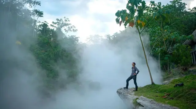 A foto mostra uma mulher na beira de uma pedra olhando para a floresta rodeada de neblina.