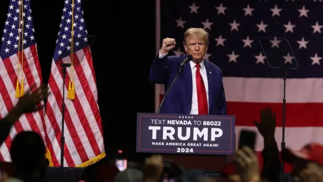 Donald Trumpo, um homem branco idoso de terno e cabelo loiro, durante um evento com apoiadores; ao fundo, bandeira dos EUA