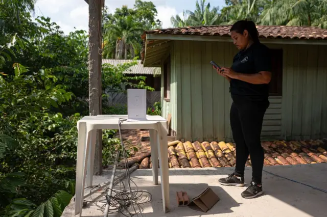 Mulher em pé em varanda, rodeada por floresta, em frente a mesa de plástico com equipamento e fios