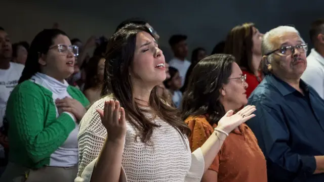 Késia ao lado dos pais em culto; ela levanta as palmas da mão para o alto e está de olhos fechados