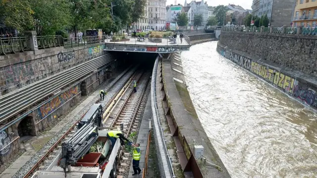 Foto de cima de um rio do lado direito e do lado esquerdo um trilho de trem onde trabalhadores retiram detritos. 