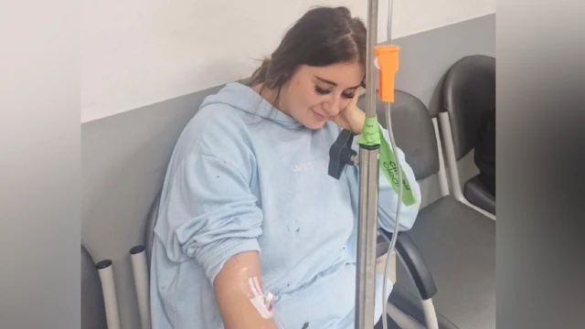 Paige Roberts sentada em uma cadeira de hospital recebendo fluidos intravenosos. Ela está vestindo uma bata de hospital azul clara e está olhando para o chão