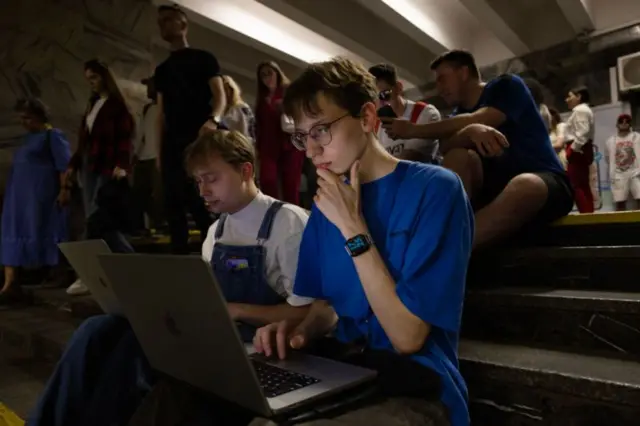 Dois jovens trabalhando em seus laptops dentro de um abrigo antiaéreo