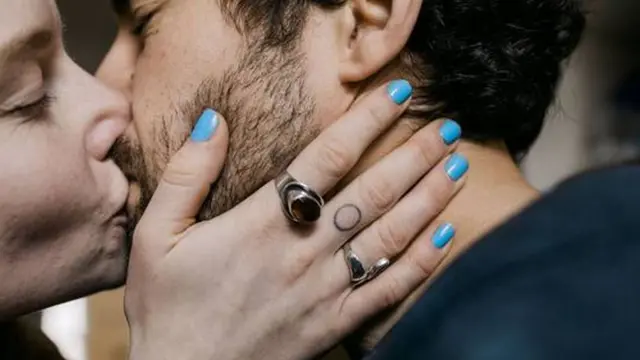 Foto colorida mostra um homem e um mulher brancos se beijando. Ele tem barba e cabelos pretos e ela segura o rosto dele com as mãos com unhas pintadas de azul