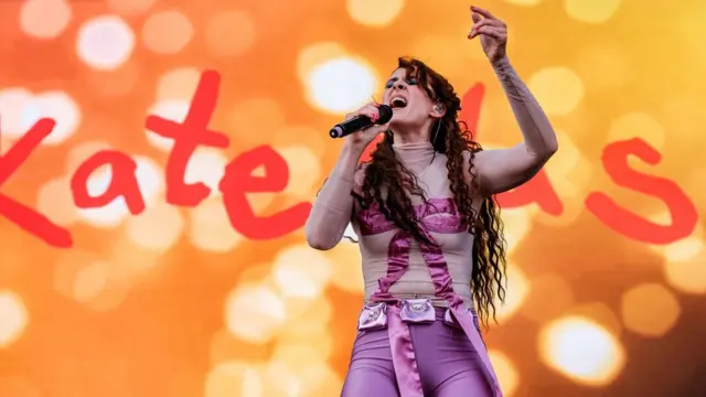 Kate Nash cantando no palco com uma roupa roxa, em um cenário laranja, com seu nome em letras gigantes