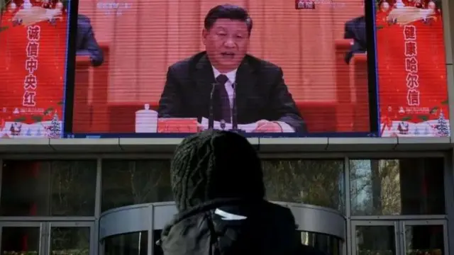 Foto de uma pessoa de costas e na frente dela, uma TV com o líder chinês Xi Jinping falando em um microfone. 