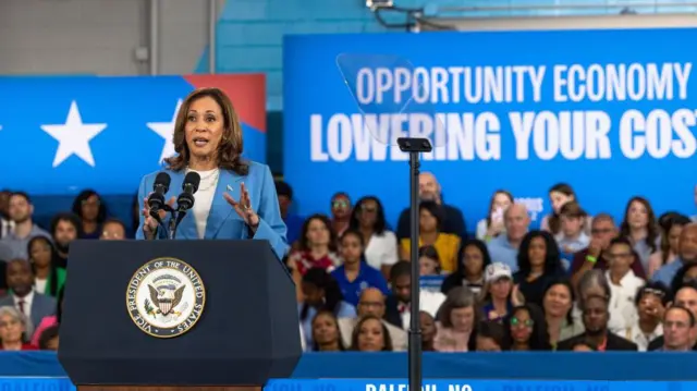 Kamala Harris discursando em palanque, com público ao fundo