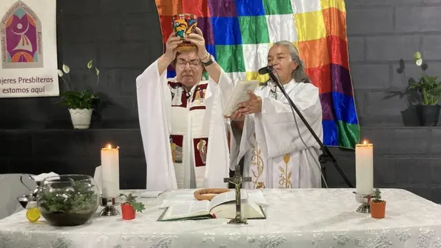 Fotografia colorida mostra mulher rezando a misso enquanto outra segura uma liturgia em sua frente