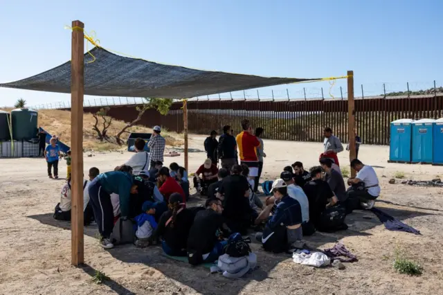 Imigrantes sentados sob tenda, próximo a banheiros químicos