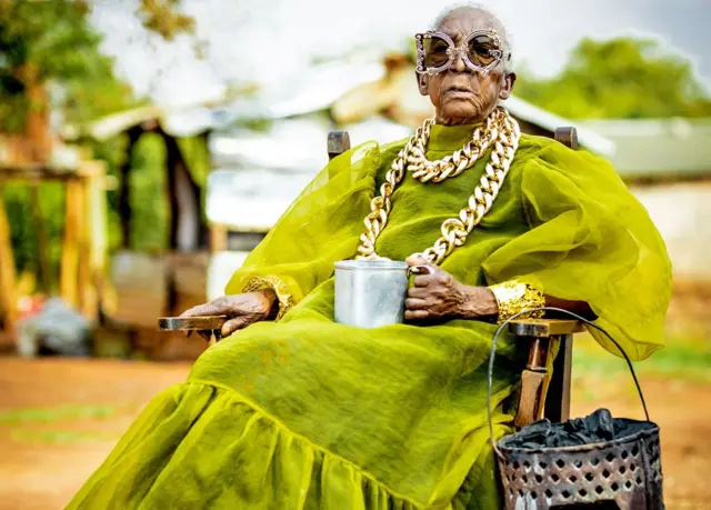 Margret Chola, sentada, usa uma corrente dourada grossa sobre um vestido verde de tafetá com mangas bufantes e uma grande babado na barra. Ela segura uma caneca de lata na mão. Pendurado no braço da cadeira, está um "mbaula" cheio de carvão. Ela usa óculos de sol com armação ornamentada, sendo que a armação do olho esquerdo tem o formato de um "D" e a do olho direito tem o formato de um "C".