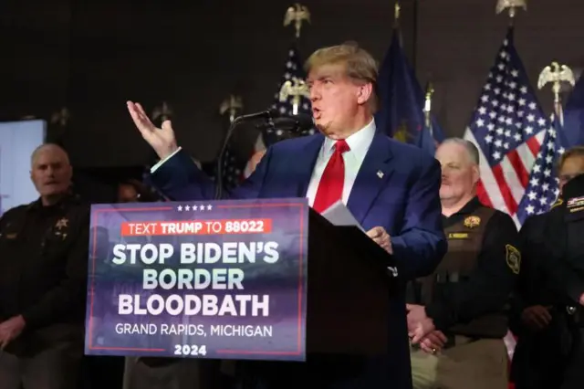 Trump discursando; abaixo dele, cartaz com slogan 'Vamos parar com o banho de sangue de Biden na fronteira'